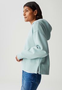 Donna con felpa con cappuccio azzurro chiaro e jeans blu, in piedi di profilo su uno sfondo semplice.