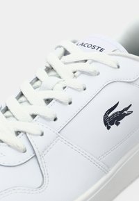 Sapatilha de couro branco com biqueira arredondada, costura tonal e um logótipo em crocodilo preto na lateral. Apresenta cadarços brancos e um colar acolchoado.