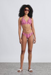 Rózsaszín, piros és fehér csíkos bikini; háromszög alakú felsővel és alacsony derekú alsóval. Fehér szandállal és négyzet alakú napszemüveggel viselve.