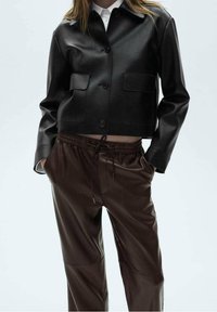 Veste courte en cuir noir avec deux poches avant, associée à un pantalon marron en simili cuir doté d'une taille élastique et d'un cordon de serrage.