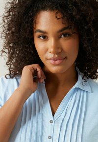 Ljusblå skjorta med knappar, plisseringar, krage och silverknappar. Tyget verkar mjukt och texturerat, vilket ger ett avslappnat utseende.