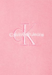 Rosa bomullstyg har broderad vit text: "Calvin Klein Jeans" med en framträdande "CK"-logotyp i en något mörkare rosa.