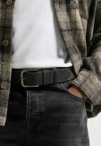 Schwarzer Stoffgürtel mit einer Metallschnalle, getragen über dunklen Jeans und einem weißen Hemd, teilweise verdeckt von einem gemusterten Flanellhemd.