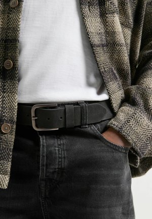 Schwarzer Stoffgürtel mit einer Metallschnalle, getragen über dunklen Jeans und einem weißen Hemd, teilweise verdeckt von einem gemusterten Flanellhemd.