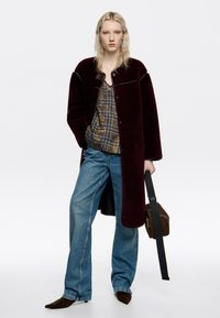 Abrigo burdeos de gran tamaño en tejido texturizado, superpuesto sobre una blusa con estampado y combinado con jeans de pierna ancha azules y accesorios marrones.