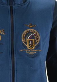 Giacca zip-up blu navy con maniche nere, caratterizzata da loghi e testi ricamati in oro e multicolore, che rappresentano una squadra di aerobatica.