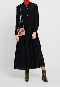 Woman wearing a long black buttoned dress, orange turtleneck, olive green patent boots, and a small beige floral handbag with a chain strap.