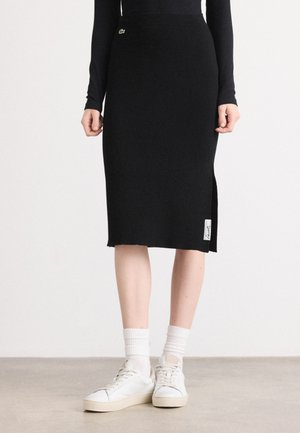 Jupe midi noire en tricot avec fente sur le côté et texture tissée, présentant un petit logo sur la taille et une étiquette en bas.