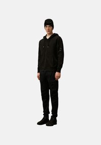 Zwarte zip-up hoodie met trekkoorden en zijzakken, gecombineerd met zwarte cargobroek met meerdere zakken en accenten. Zwarte beanie op.