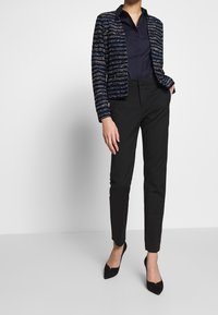 Femme portant un pantalon noir, des talons aiguilles noirs, un blouse marine et une veste texturée bleu et blanche à rayures, se tenant avec une main dans la poche.