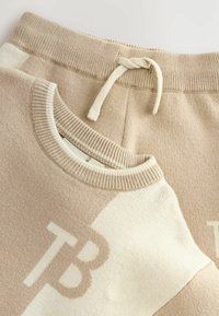 Beige sweater en bijpassende sweatpants, met een gebreide textuur. Accenten zijn een trekkoord en een kleurblokontwerp met logo-detail.