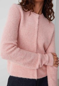 Cardigan rose clair en matériau côtelé doux, avec un devant à boutons et des manches longues dotées de poignet légèrement ajustés.