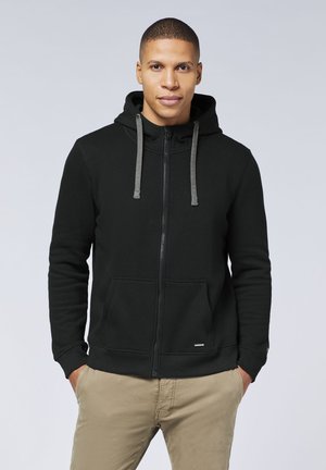 Schwarzer Reißverschluss-Hoodie mit grauen Kordeln, Fronttaschen und einer glatten Textur. Trägt dazu khakifarbene Hosen, mit einem Logo am unteren Saum.
