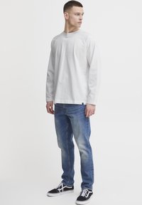 Langärmliges, Rundhals weißes Baumwoll-T-Shirt kombiniert mit hellblauen Jeans; Model steht in schwarzen Sneakers mit weißen Sohlen.
