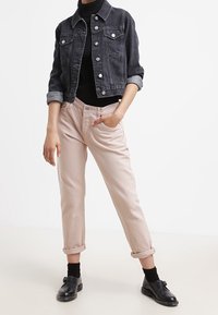 Chaqueta de mezclilla negra con botones plateados, parte superior negra, pantalones rosa pálido con puños enrollados y zapatos negros con detalle de correa.