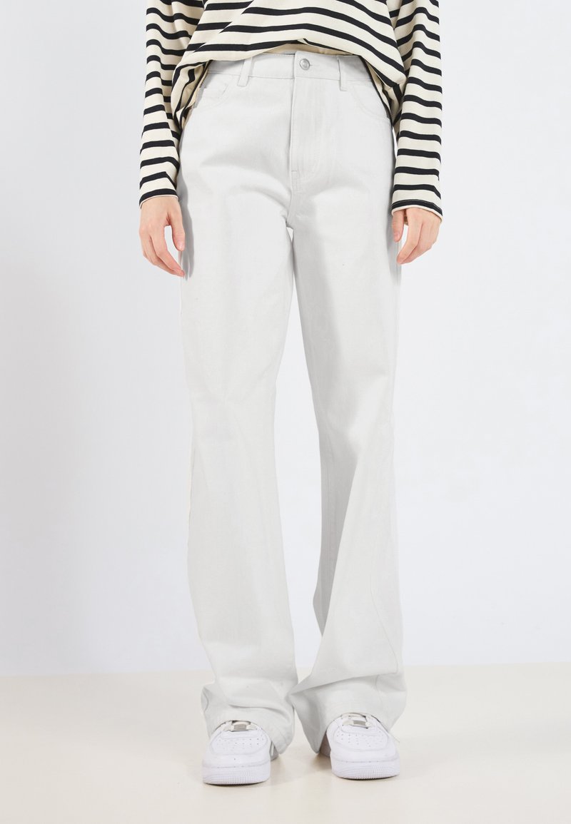 Calças largas de cor cinza-claro, feitas de um tecido macio com uma textura suave. Combinadas com uma blusa listrada em preto e bege e ténis brancos.