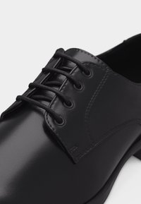 Μαύρο δερμάτινο παπούτσι με κλειστό κορδόνι, λείο γυαλιστερό ύφασμα και στρογγυλά μαύρα κορδόνια δεμένα μέσω τεσσάρων οπών σε κάθε πλευρά.