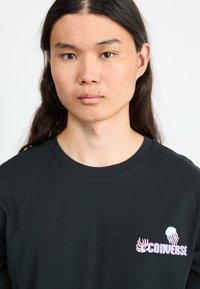 Sort bomulds T-shirt med rund hals. Har et trykt logo med en kvalmotivet i lilla og hvidt på venstre brystområde.