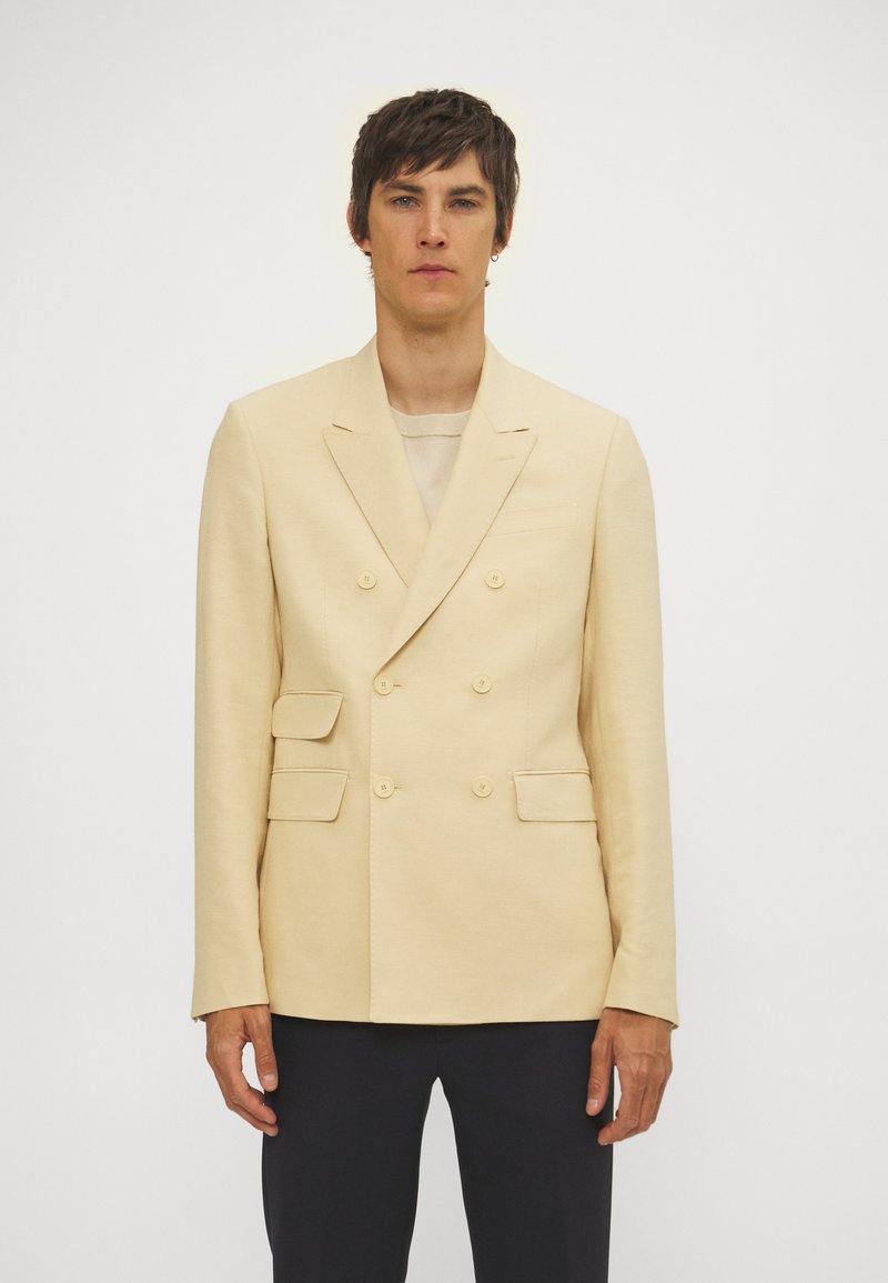 Beige dubbelzijdige blazer met twee voorzakken en gouden knopen. Beschikt over gekartelde revers en een gladde stoftextuur.