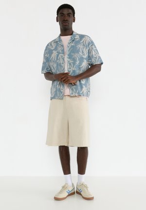 Kurzärmeliges, blau gemustertes Hemd über einem hellrosa T-Shirt, kombiniert mit beigen Shorts und cremefarbigen Sneakern mit blauen Akzenten. Schlichte, lockere Passform.