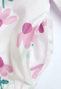 Tessuto con motivo floreale in colori rosa e verde, caratterizzato da una texture leggera e un dettaglio con apertura laterale. Sfondo bianco morbido.