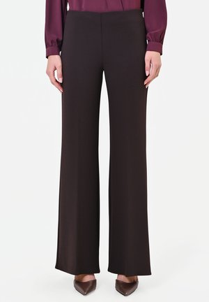 Donna con pantaloni neri a gamba larga, una camicetta bordeaux a maniche lunghe e scarpe marroni con punta, in piedi davanti a uno sfondo bianco.