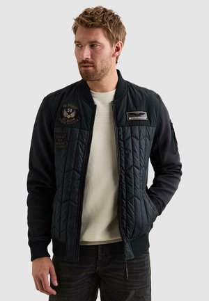 Homme aux cheveux châtain clair et à la barbe portant une veste bomber matelassée noire avec des écussons et un pull en maille blanc, regardant sur le côté.