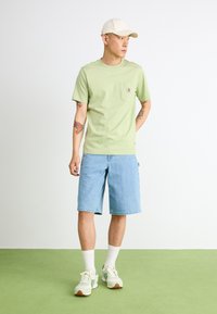 T-shirt mint vert à manches courtes avec une petite poche poitrine, associée à un short en denim bleu clair. Les accessoires comprennent une casquette beige et des baskets légères.