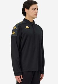 Camiseta atlética negra de manga larga con cuello de media cremallera, que presenta acentos de logo dorado en los hombros y una tela suave y transpirable.