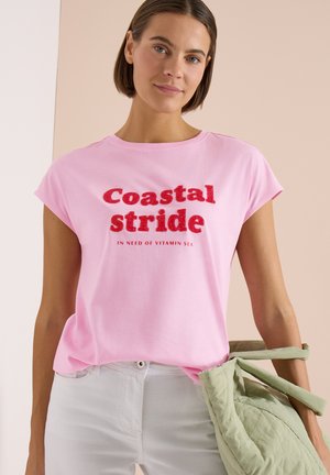 Frau trägt ein hellrosa Shirt mit rotem Text „Coastal stride in need of vitamin sea“ und hält eine hellgrüne Tragetasche am Arm.