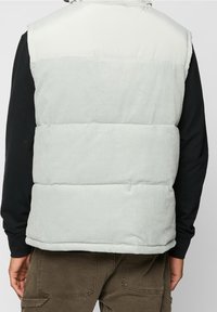 Man draagt een lichtgrijs, gewatteerd mouwloos jack over een zwarte lange mouw en bruine broek, rug naar de camera gericht.