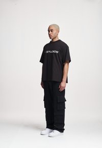 T-shirt noire surdimensionnée avec "STUDIOS" en lettres blanches, associée à un pantalon cargo noir avec des poches et des baskets blanches.