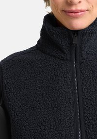 Schwarze Fleece-Weste mit strukturierter Oberfläche, hohem Kragen und Reißverschluss. Besitzt schwarze Materialeinsätze an den Seiten für einen Kontrast.