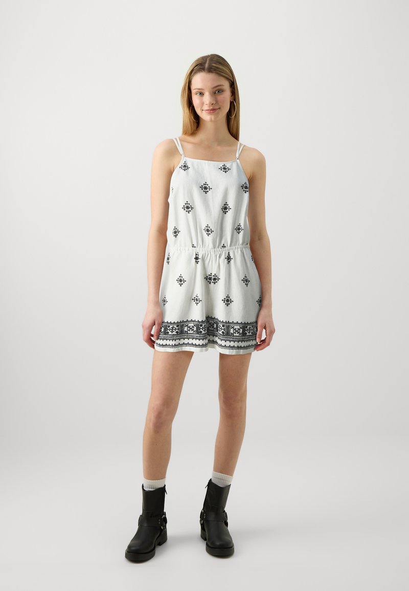 Vestido sin mangas blanco con patrones geométricos negros, cinturilla elástica y borde de encaje en el dobladillo. Llevado con botas negras hasta el tobillo y calcetines blancos.