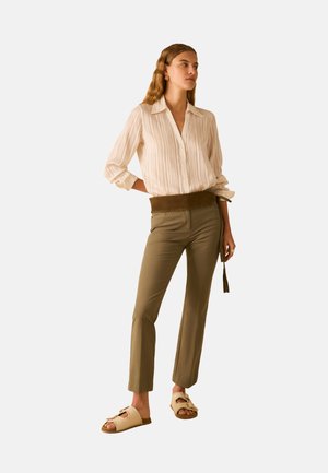 Donna che indossa una blusa beige a righe, pantaloni slim verde oliva con una cintura larga e sandali beige intrecciati con punta aperta, in piedi con una mano dietro la schiena.