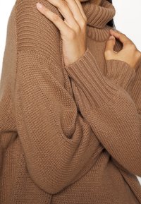 Brązowy sweter z dzianiny z wysokim golfem, z prążkowanymi mankietami i różnicami w fakturze materiału.