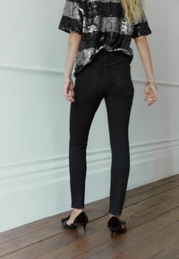 Čierne džínsy s vysokým pásom a slim fit, v kombinácii s topom posiatym flitrami. Oblečenie dopĺňajú čierne lodičky s úzkou špičkou a drevená podlaha v pozadí.
