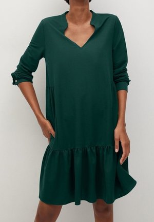Frau trägt ein dunkelgrünes, langärmliges Kleid mit V-förmigem Ausschnitt und gerüschtem Saum, steht mit einer Hand in der Tasche.