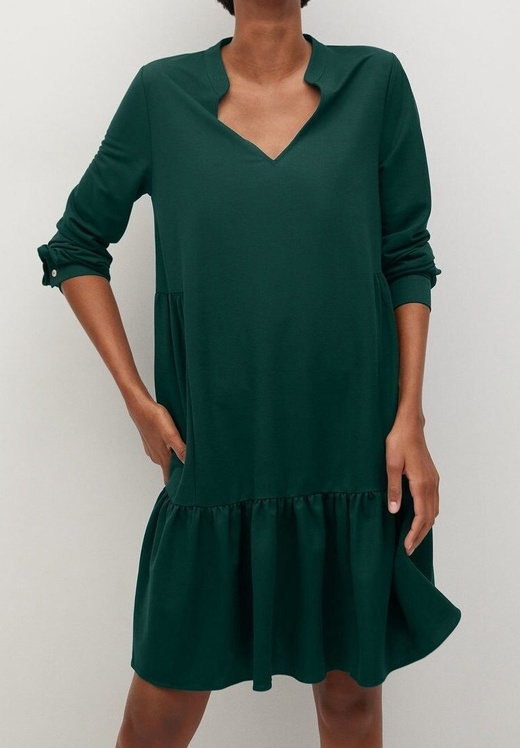 Femme portant une robe à manches longues vert foncé avec un col en V entaillé et un ourlet à volants, debout avec une main dans la poche.