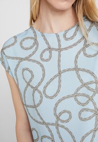 Top azul claro hecho de una tela suave con un patrón de cadenas en marrón y dorado. Tiene mangas cortas y un escote redondeado.