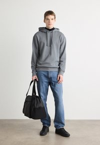 Grijze hoodie met een voorzak, gecombineerd met blauwe jeans. Het model houdt een zwarte sporttas vast en draagt zwarte sneakers. Minimalistisch ontwerp.