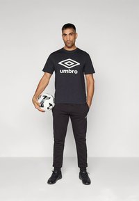 Zwart T-shirt met korte mouwen en wit "Umbro"-logo, gecombineerd met zwarte joggingbroek en zwarte sportschoenen, terwijl een voetbal met patroon wordt vastgehouden.