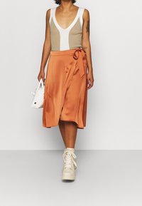 Top a canotta a coste di colore beige con scollatura a V profonda, abbinato a una gonna avvolgente in satin arancione con un nodo in vita e orlo a ginocchio. Sneakers bianche.