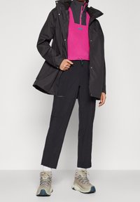 Veste imperméable noire portée sur un haut en polaire rose, associée à un pantalon noir et des bottes de randonnée beige. Comprend des détails avec des fermetures éclair et un cordon de serrage.