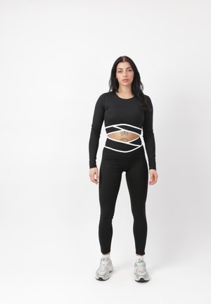 Vrouw staand in een zwarte cropped top met lange mouwen en legging met witte accenten, toont een buik-tatoeage, draagt witte sneakers op een witte achtergrond.