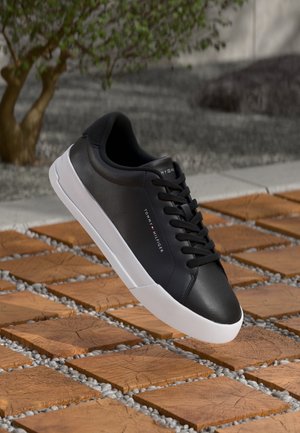 Sort læder sneaker med hvid sål og sorte snørebånd, "Tommy Hilfiger" branding, vist over træfliser og grus ude.