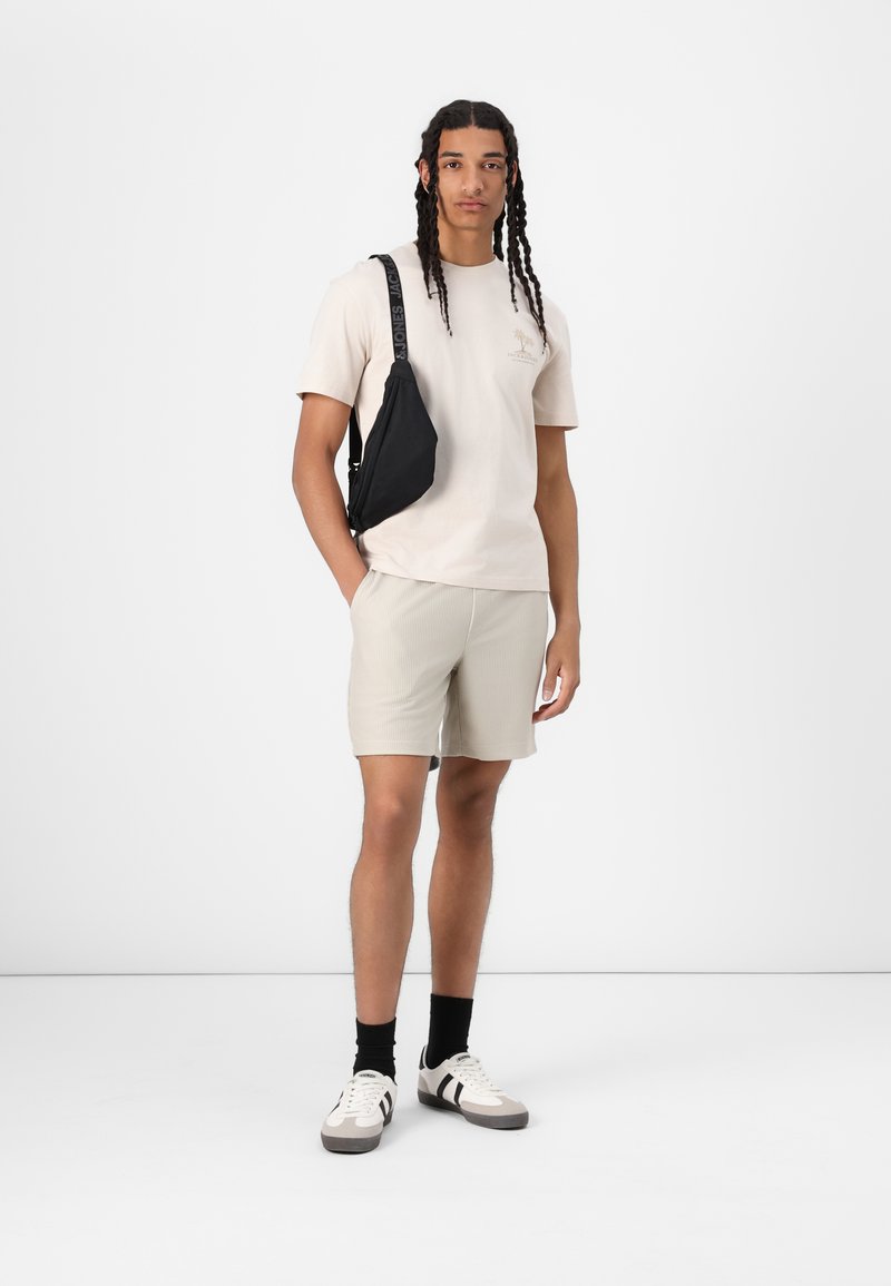 Beige t-shirt with logo, light gray ribbed shorts, black crossbody bag, black socks, and two-tone sneakers. Plain white background.