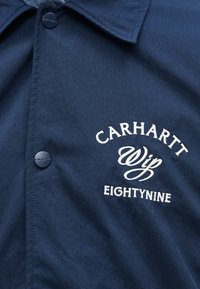 Veste Carhartt bleu marine avec boutons-pression et texte brodé blanc "CARHARTT WIP EIGHTYNINE" sur la poitrine.