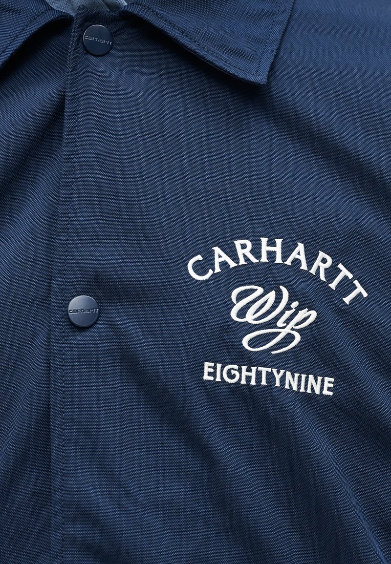Giacca Carhartt blu navy con bottoni a pressione e testo ricamato bianco "CARHARTT WIP EIGHTYNINE" sul petto.