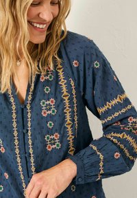 Blaue Stoffbluse mit gestickten Mustern in Gelb, Grün und Rot. Mit Knöpfen auf der Vorderseite und gefältelten Ärmelbündchen.